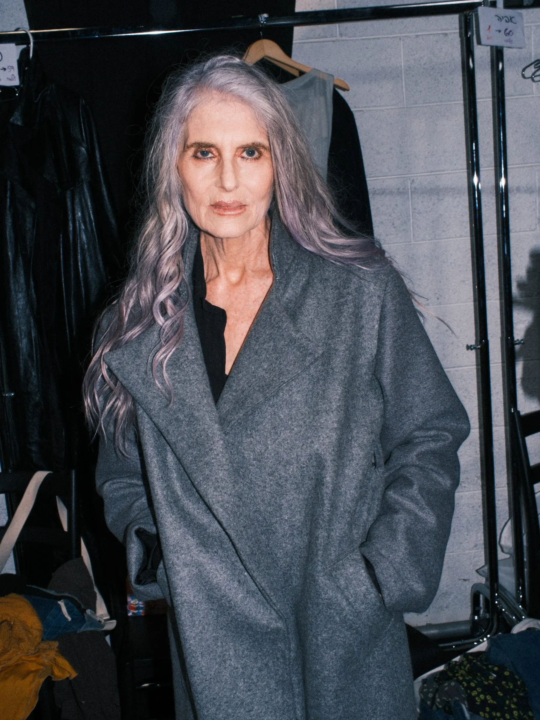 Model with long silver hair in an oversized grey wool coat, backstage beside clothing rails