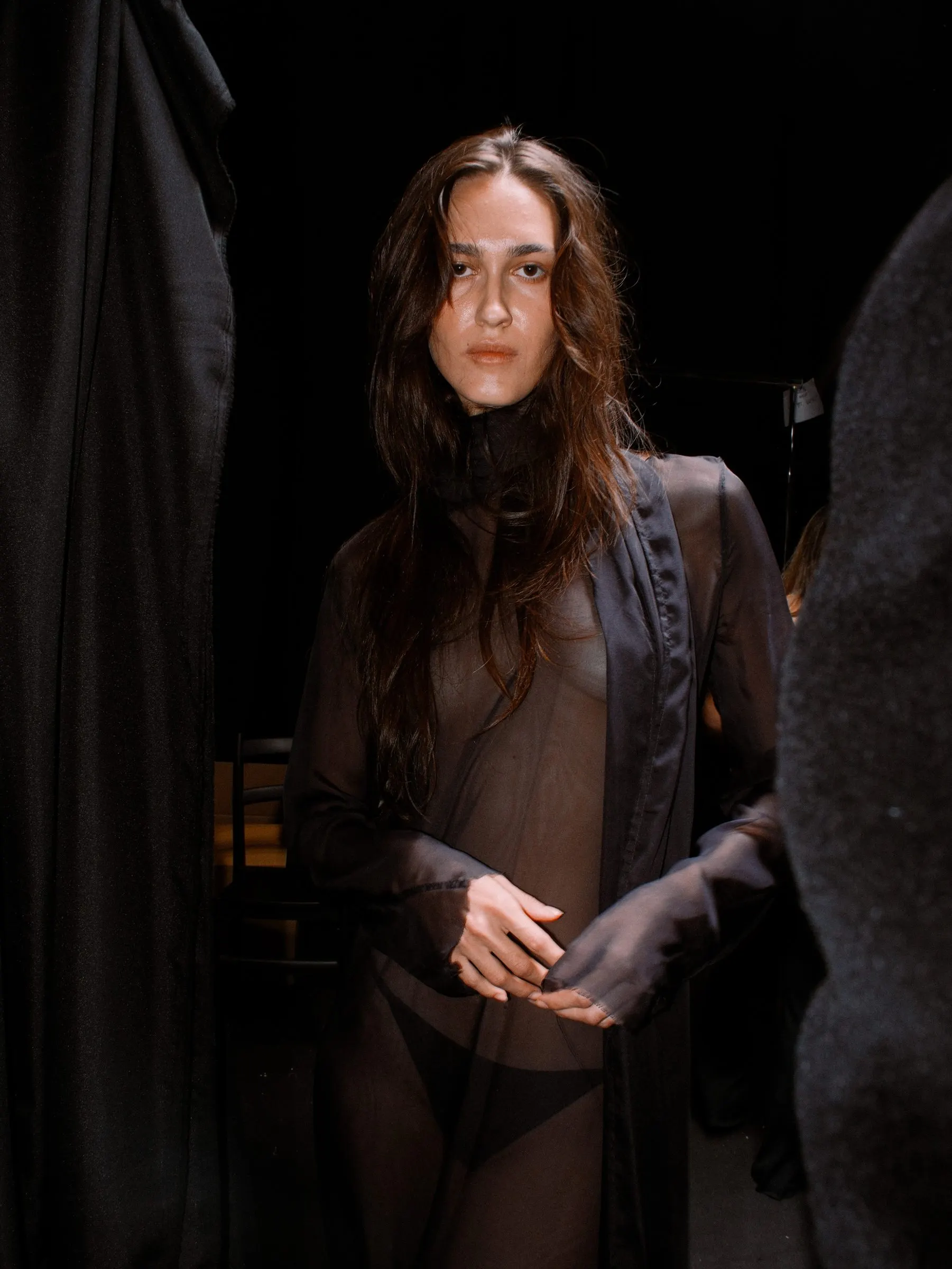 Sheer black layered look under a tailored jacket, hands clasped against a dark curtain