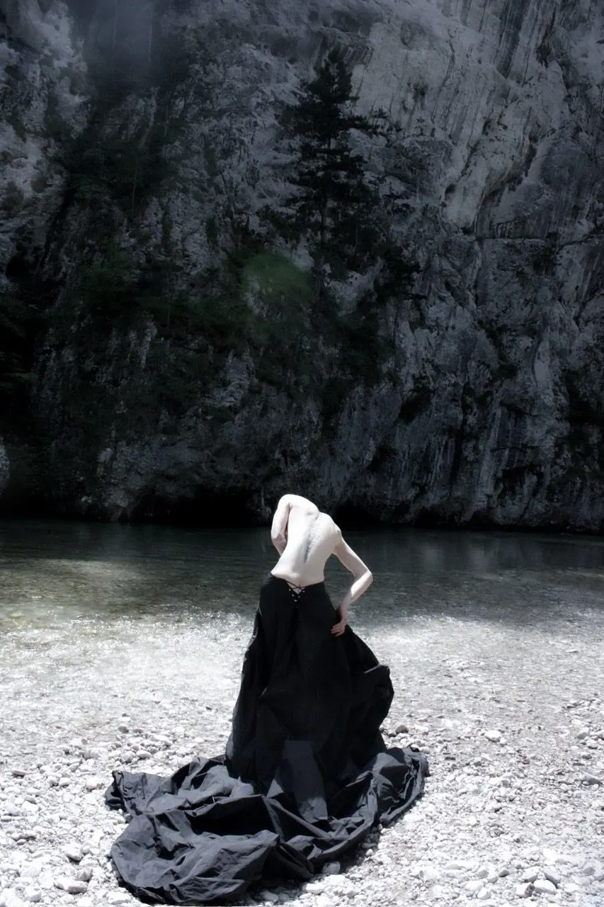 Voluminous black drapery on a rocky riverbank, the figure turned away with head bowed