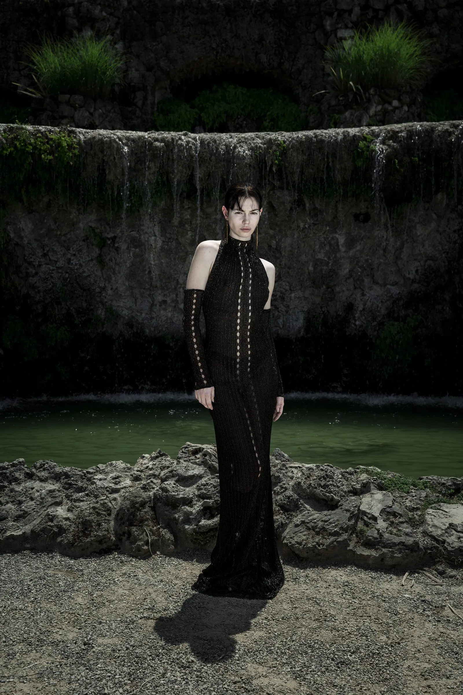 Long black column dress with laced sleeves, shot before a mossy stone wall and water