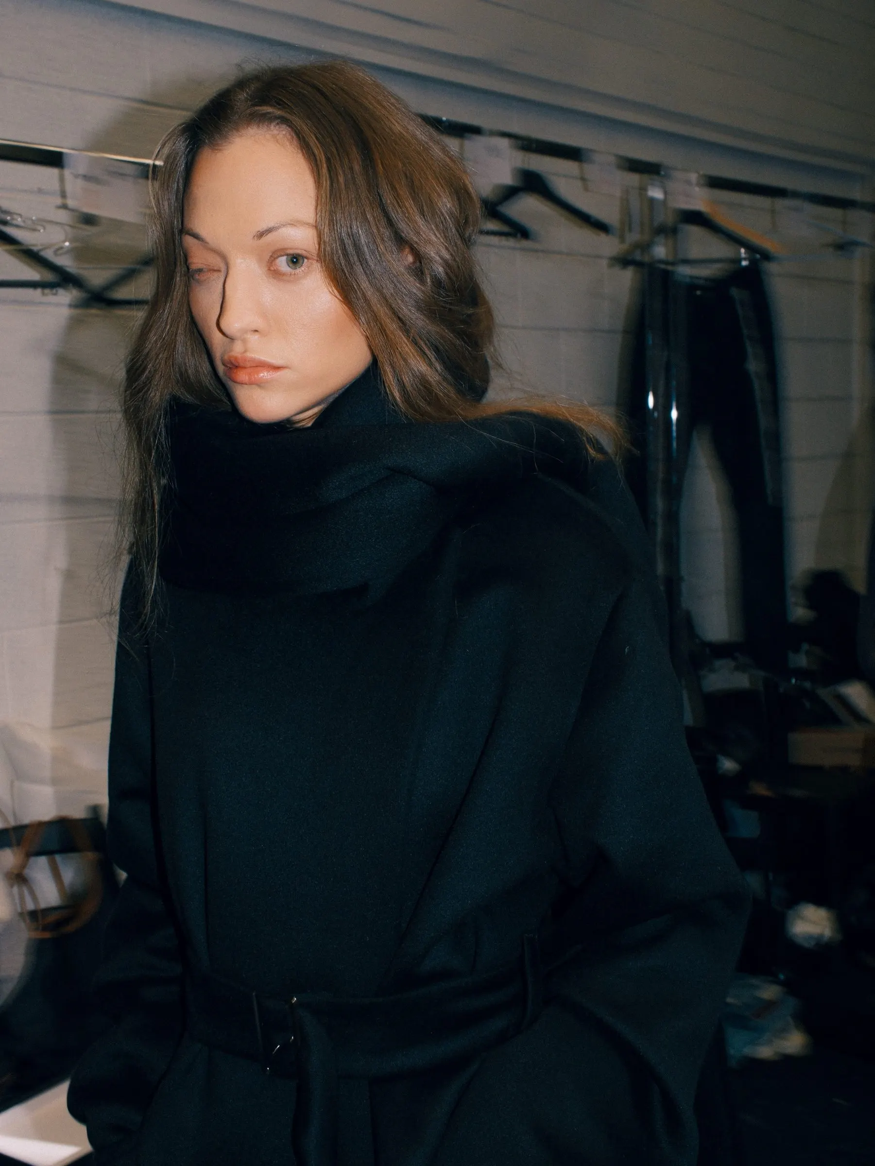 Black coat with a wide upturned collar, worn backstage in front of garment rails
