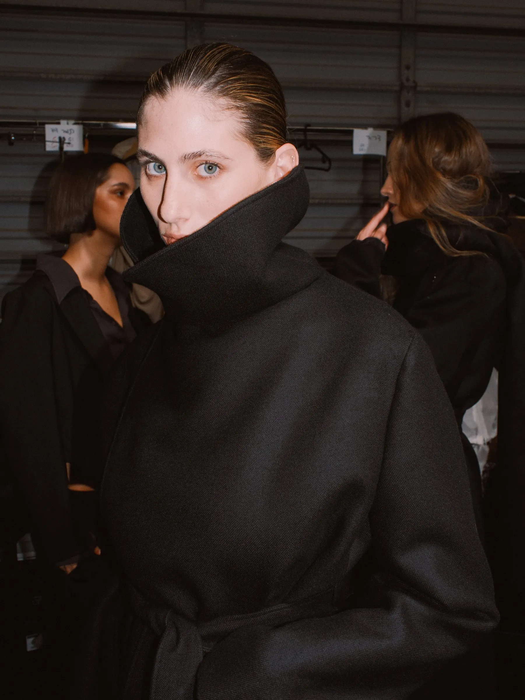 Oversized black wool coat with the collar turned up, backstage among other looks