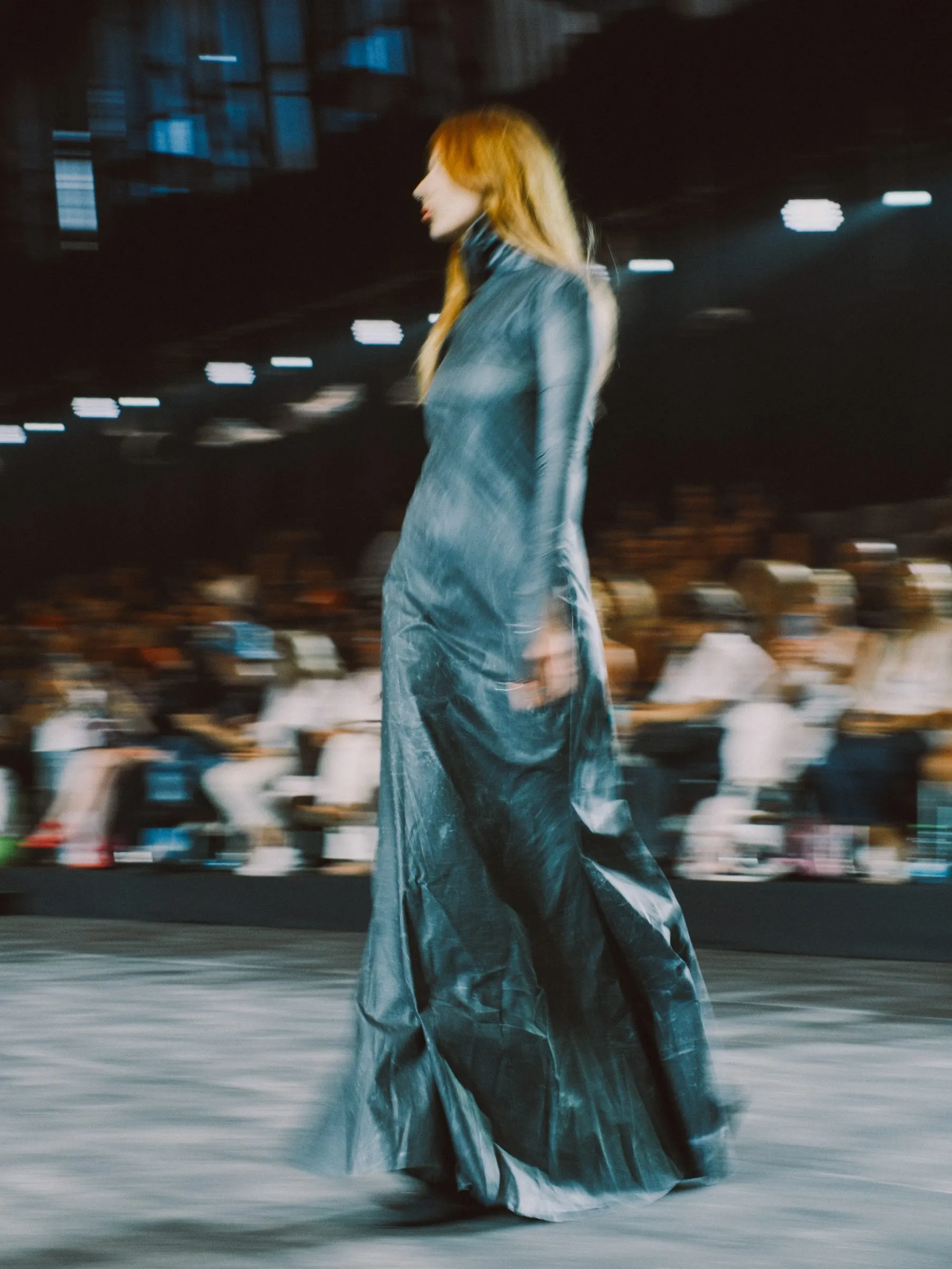 Runway shot in motion blur: a floor-length draped gown in teal, audience seated behind